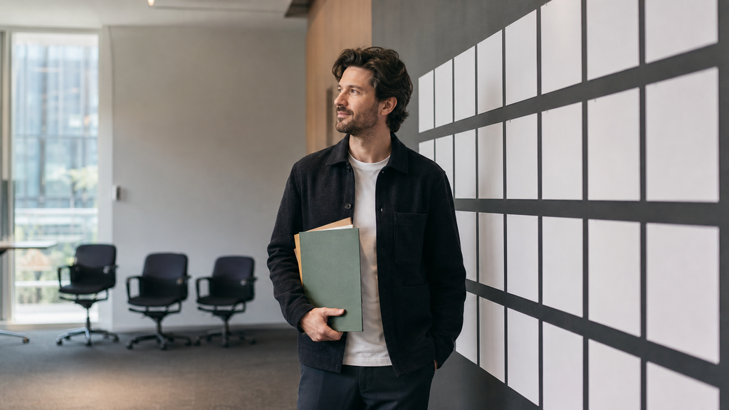 Founder holding renewal folders beside a planning wall, representing converting monthly SaaS customers to annual contracts before a sale