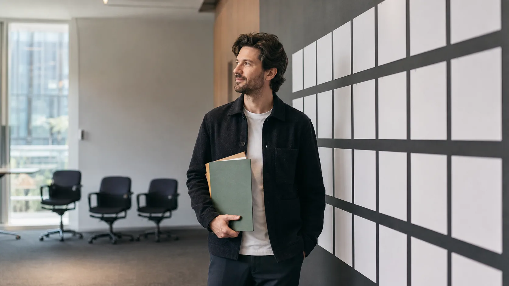 Founder holding renewal folders beside a planning wall, representing converting monthly SaaS customers to annual contracts before a sale